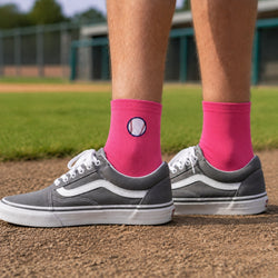 A person standing on a baseball field wearing gray sneakers and bright pink crew socks with a baseball graphic on the cuff.