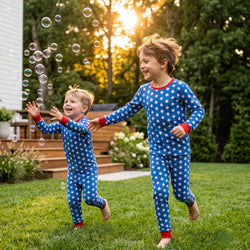 Two young children running and playing on a green lawn in a backyard at sunset, wearing matching blue long-sleeve pajamas with white baseball pattern and red trim.