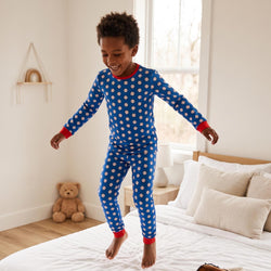 A young boy wearing a blue long-sleeve pajama set with white baseball patterns and red trim, jumping excitedly on a white bed in a bright bedroom.