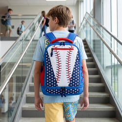 A boy wearing a baseball-themed backpack stands on an indoor staircase, viewed from behind. The backpack is bright blue with white polka dots, a white center panel, and red seam details, matching a baseball design.