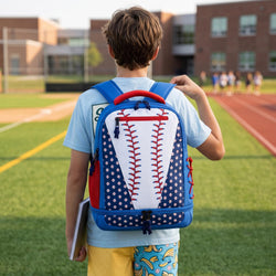 A child viewed from behind wearing a baseball-themed backpack on a school track field. The backpack is blue and white with red stitching details, and the child is wearing a light blue t-shirt and yellow pants.