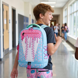 A smiling boy in a school hallway wearing a light blue and pink backpack with a baseball stitching design, pink dripping paint details, and a green top handle.