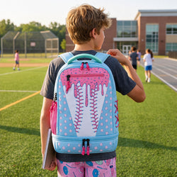 A child viewed from behind on a school field, wearing a light blue and pink baseball-themed backpack with a dripping pink design and white baseball stitching graphic.