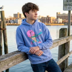 Person wearing a blue hoodie with a cartoon ice cream baseball character on a dock by water