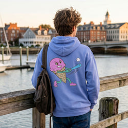 Person wearing a blue hoodie with a colorful baseball ice cream graphic, standing by a waterfront with buildings and water in the background.