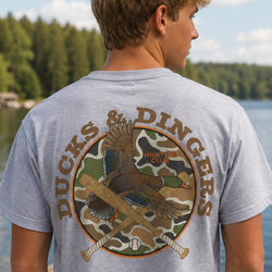 A man wearing a heather gray t-shirt with a large back graphic that reads 'DUCKS & DINGERS HUNTING CLUB' around a duck, baseball, and crossed bats over a camouflage circle, photographed outdoors by a lake.