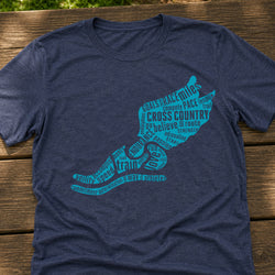 A navy blue heathered t-shirt laid flat on a rustic wooden surface, featuring a graphic of winged run shoe filled with running and cross-country training words in bright teal.