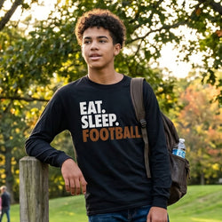 Person wearing a black long sleeve shirt with 'EAT. SLEEP. FOOTBALL' text in an outdoor setting.
