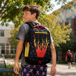 A boy wearing a gray t-shirt and black shorts walks outdoors on a campus-style path while wearing a black backpack with a red, orange, and yellow football flame design on the front.