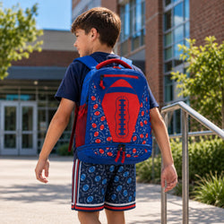 A child viewed from behind wearing a blue and red football-themed backpack on an outdoor sports field, with other children and footballs blurred in the background.
