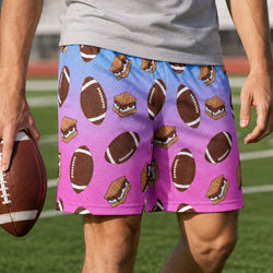 A person wearing mesh athletic shorts with a blue-to-pink gradient background and an all-over football and s'mores print, standing on a grassy field and holding a football.