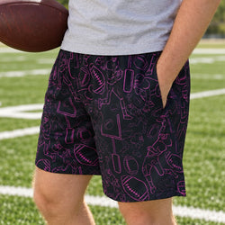 A man wearing black athletic shorts with a pink football-themed pattern, standing on a green football field and holding a brown football.