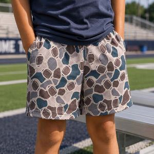A person standing on a sports field wearing patterned shorts with a navy blue top. The shorts feature an abstract football spot pattern in shades of blue, brown, and gray on a light beige background.