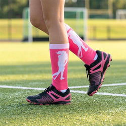 Person wearing pink socks with a white field hockey design and black cleats on a grass field.