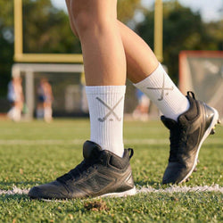 Person wearing black shoes and white socks with a field hockey logo on a grassy field.