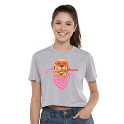 A smiling young woman wearing a light heather gray cropped T-shirt with a graphic of a dog wearing orange sunglasses and a pink bandana, holding a pink field hockey stick. She is also wearing blue jeans against a plain white studio background.
