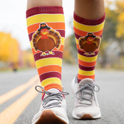 A person wearing mid-calf Thanksgiving-themed striped socks with a turkey graphic, shown from the knees down on an outdoor road with blurred autumn trees in the background.