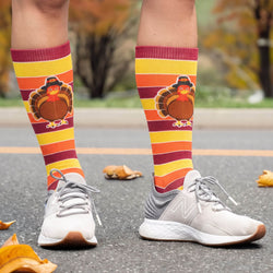 A person wearing mid-calf Thanksgiving-themed socks with a turkey graphic and orange, yellow, and burgundy stripes, paired with light gray sneakers on an outdoor paved path with scattered autumn leaves.