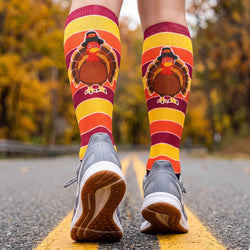 A person running on an asphalt road wearing knee-high socks with a large turkey graphic and bold horizontal stripes in yellow, orange, red, and burgundy, paired with gray sneakers.