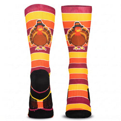A pair of crew socks with a Thanksgiving theme, featuring a cartoon turkey wearing a pilgrim hat on a background of horizontal orange, yellow, and burgundy stripes, with a black foot section.