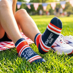 Person wearing red, white, and blue ankle socks with Land That I Love, Home Sweet Home text sitting on grass