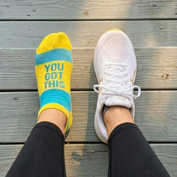 White sneaker and yellow socks with 'You Got This' text on a wooden floor.