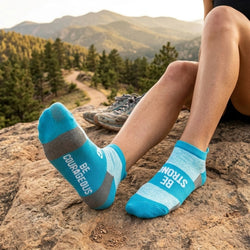 Person wearing blue socks with motivational Be Strong, Be Courageous text in a mountainous landscape