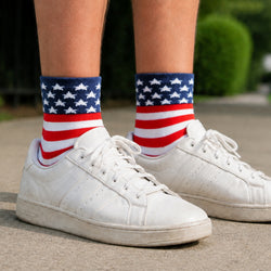 A close-up of a person's legs wearing white sneakers and patriotic crew socks with a blue star pattern and red and white stripes, standing on a paved outdoor surface.