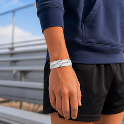 A close-up of a person wearing a white wristband with black dotted lines, paired with a navy blue hoodie and black athletic shorts, standing outdoors near metal bleachers.