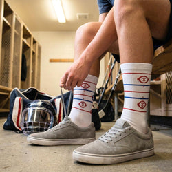 Person sitting in a locker room wearing gray sneakers and white socks with red and blue hockey rink design.