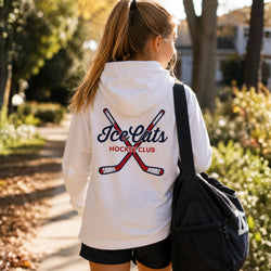 A girl seen from behind wearing a white hoodie with a 'Ice Cats Hockey Club' graphic on the back, carrying a black duffel bag on a sunny outdoor path.