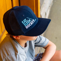 Boy wearing a navy blue mesh hockey trucker hat featuring embroidered Eat Sleep Hockey slogan and stick graphic.