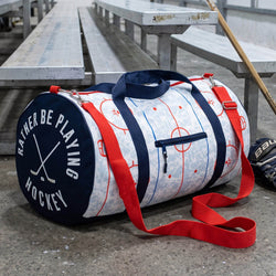 A hockey duffel bag sits on a concrete floor between metal bleachers. The bag has a white base with a light blue hockey rink pattern, navy blue handles and side straps, and bright red webbing straps and trim.