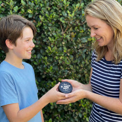 Boy giving his mom a 'Best Hockey Mom Ever' puck with green leafy background