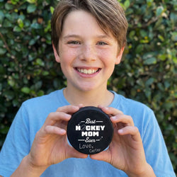 Boy holding a personalized hockey puck with 'Best Hockey Mom Ever' text against a green leafy background