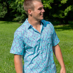 A man wearing a light blue short-sleeve button-up shirt with a red and blue hockey stick pattern, standing outdoors in a grassy park.