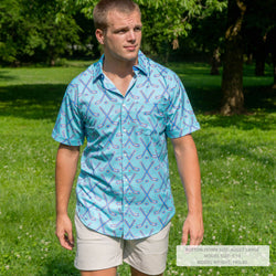 A man wearing a light blue short-sleeve button-down shirt with a repeating pattern of crossed hockey sticks in pink and purple, standing outdoors in a grassy park with trees in the background.