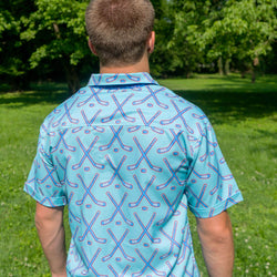 A man viewed from behind wearing a light blue short-sleeve collared shirt with a repeating pattern of crossed hockey sticks in blue and red, standing on a grassy lawn with trees in the background.