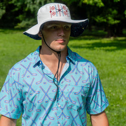 A man wearing a white bucket hat with a red and blue target pattern on the front and a blue short-sleeve button-up shirt with a hockey stick pattern, standing outdoors in a grassy park.