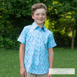 A young boy smiling outdoors in a light blue short-sleeve button-up shirt with a purple and blue hockey stick pattern, standing on a grassy lawn with trees in the background.
