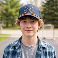 Young boy wearing a blue cap with Dangle Snipe Celly text, standing outdoors with trees in the background