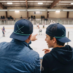 Two people wearing navy blue caps with a hockey stick design, pointing towards an ice rink.