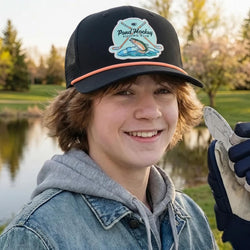 Young boy wearing a cap with a 'Pond Hockey Fishing Club' logo, holding hockey gear by a lake.