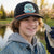 Young boy wearing a cap with a 'Pond Hockey Fishing Club' logo, holding hockey gear by a lake.