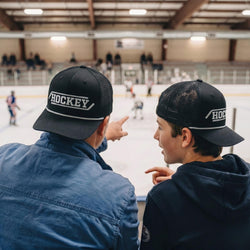 Two people wearing 'Hockey' caps at an indoor ice rink.