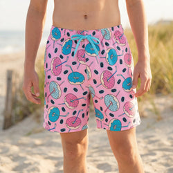 Person wearing pink swim trunks with blue and pinkhockey  donut pattern on a beach.
