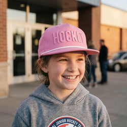 Child wearing a pink 'Hockey' cap and gray hoodie with a logo, standing outdoors.