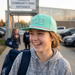 Person wearing a teal 'HOCKEY' cap with a backpack, standing in front of a building entrance.