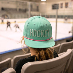 Person wearing a teal 'HOCKEY' cap at an ice hockey rink