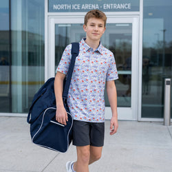 Young boy with a hockey patterned polo shirt and dark shorts holding a blue backpack outside an ice arena entrance.